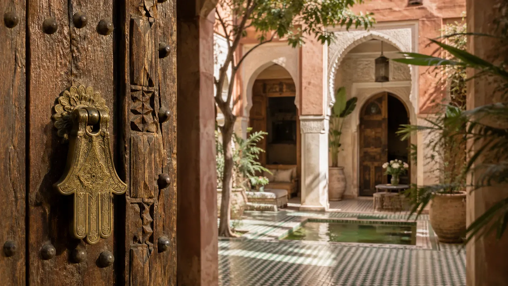 Heurtoir en laiton en forme de main de Fatma sur la porte en bois entrouverte d’un riad de Marrakech, patio à arcades en arrière-plan